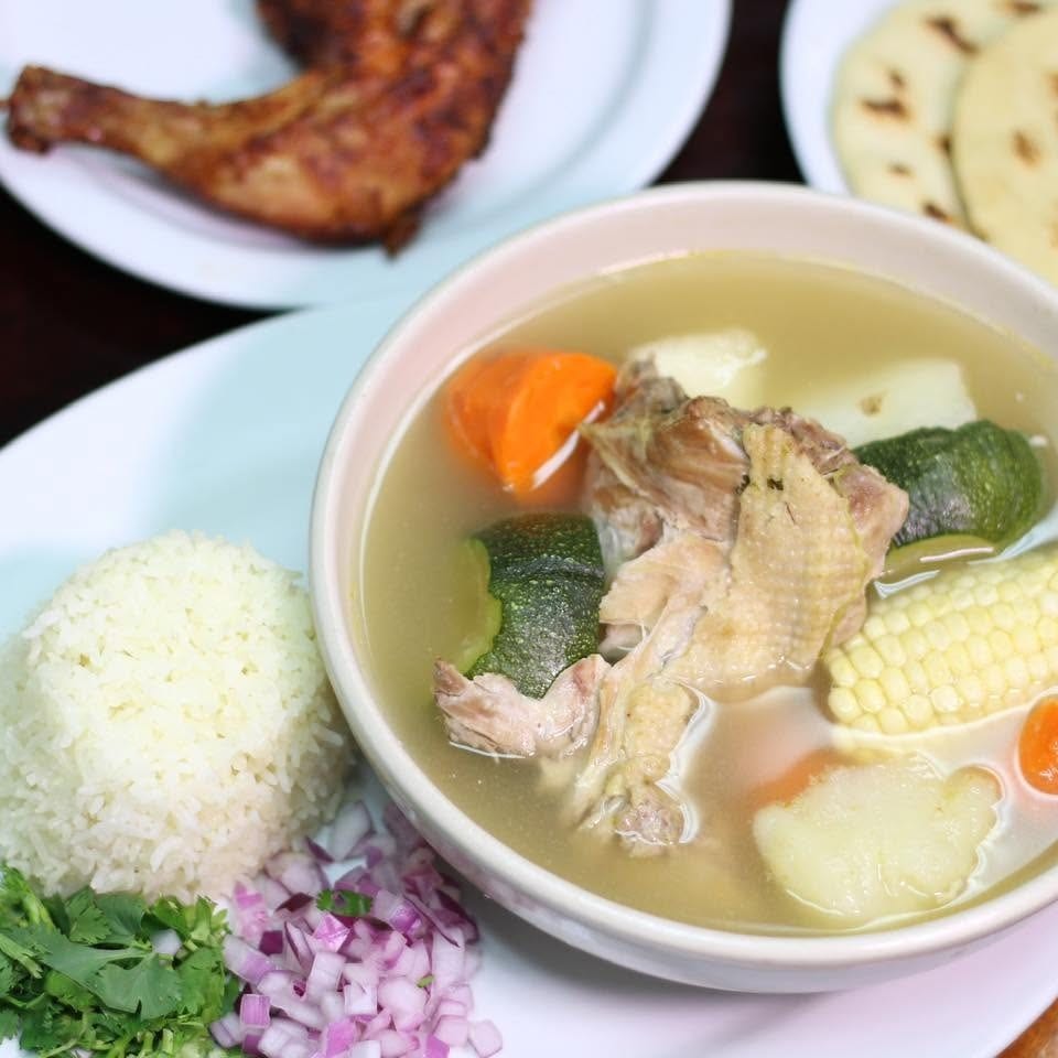 Bowl of chicken and vegetable soup served with rice, cilantro, chopped red onion, fried chicken, and flatbreads.