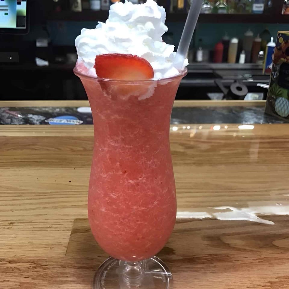 Pink frozen strawberry drink in a stemmed glass, topped with whipped cream and a strawberry, sits on a restaurant bar.