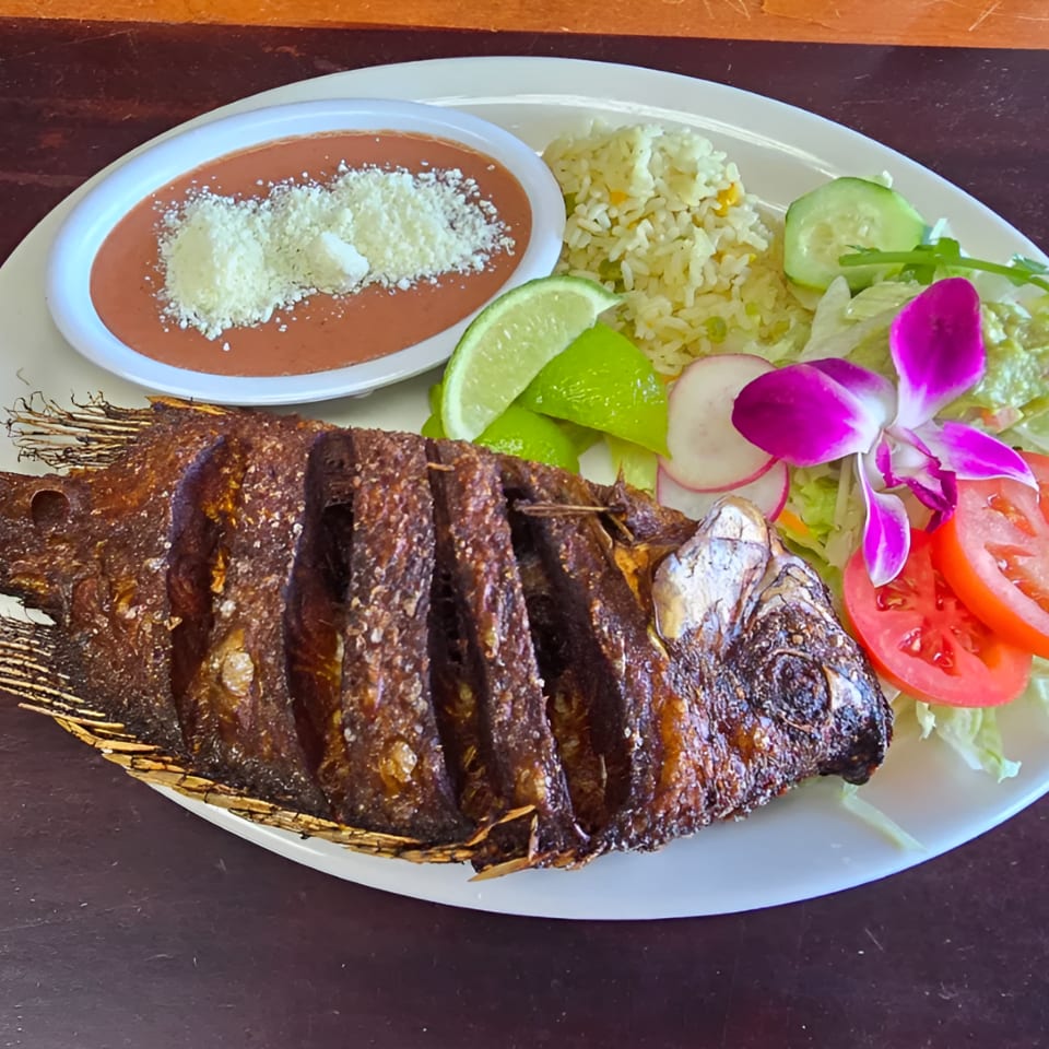 Whole grilled fish served with rice, beans topped with cheese, salad, lime wedges, and a purple orchid garnish.