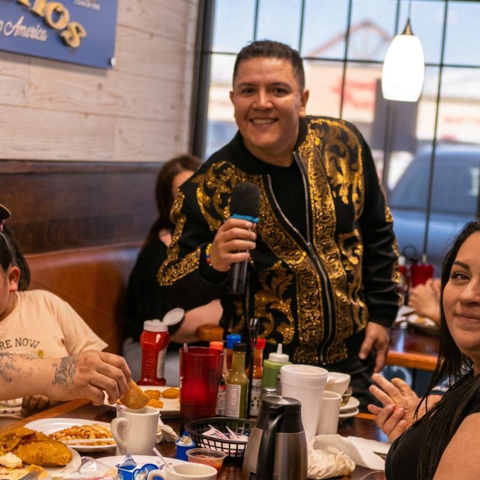 Group dining in a Central American restaurant as a man in an ornate jacket speaks into a microphone beside a table of food.