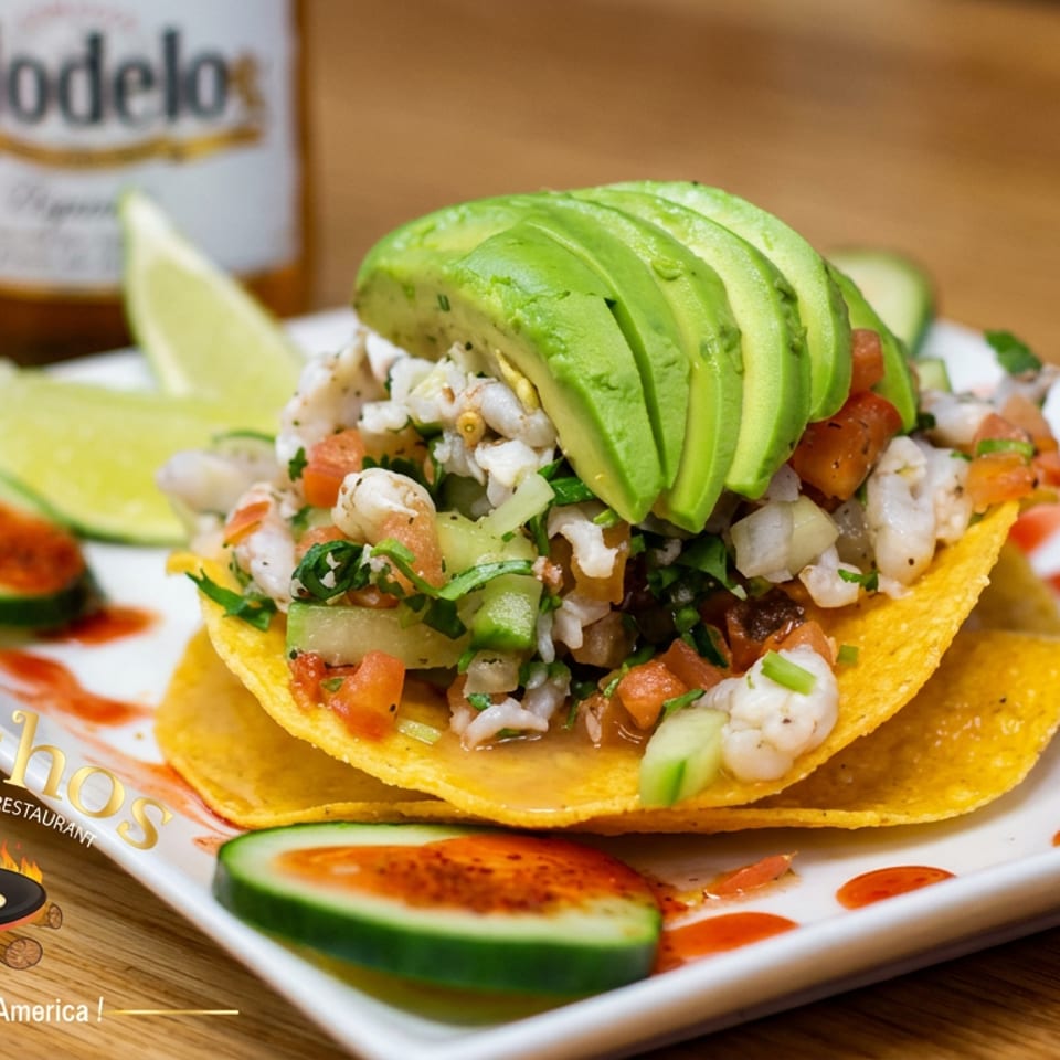 Seafood tostada topped with sliced avocado, pico de gallo, lime wedges, and chili-seasoned cucumber slices on a plate.