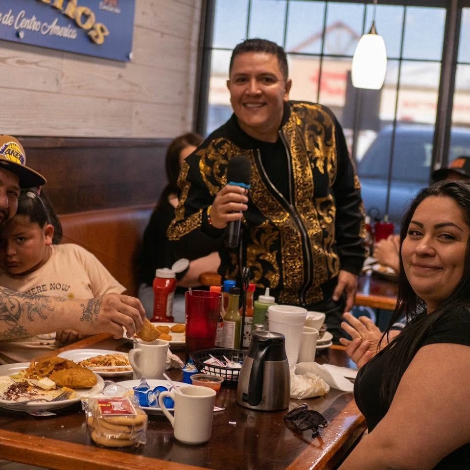Family and friends gather around a restaurant table with Latin American dishes as a smiling man holds a microphone.