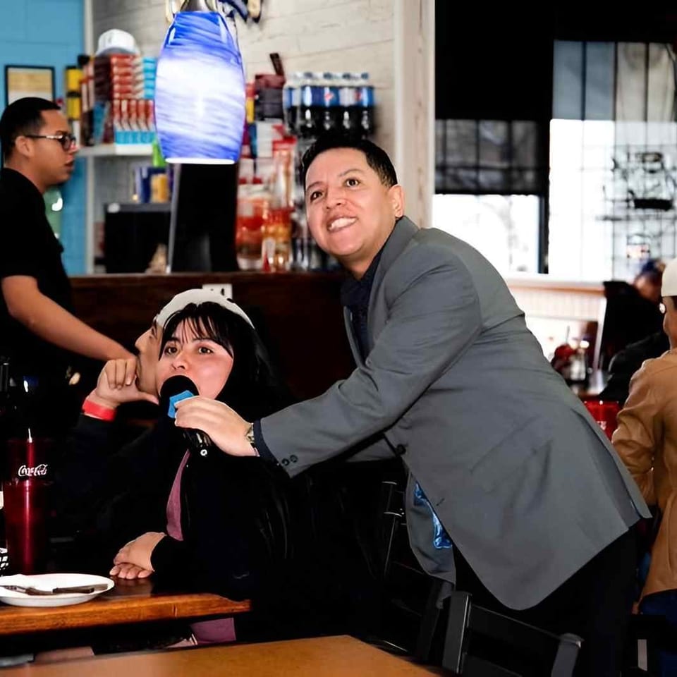 Restaurant scene with a smiling man holding a microphone toward a seated woman, surrounded by diners and colorful pendant lights.