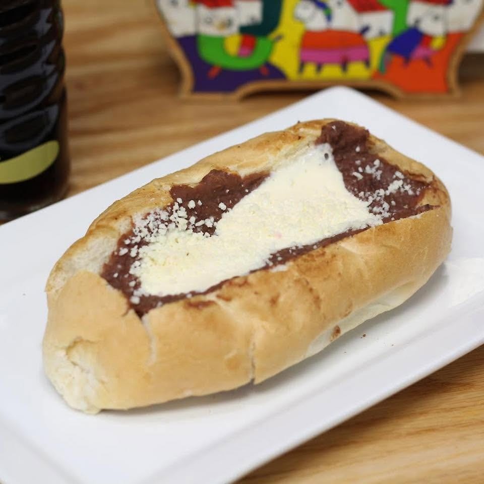 Bolillo pastry topped with chocolate spread and crumbled white cheese on a plate beside a dark beverage.