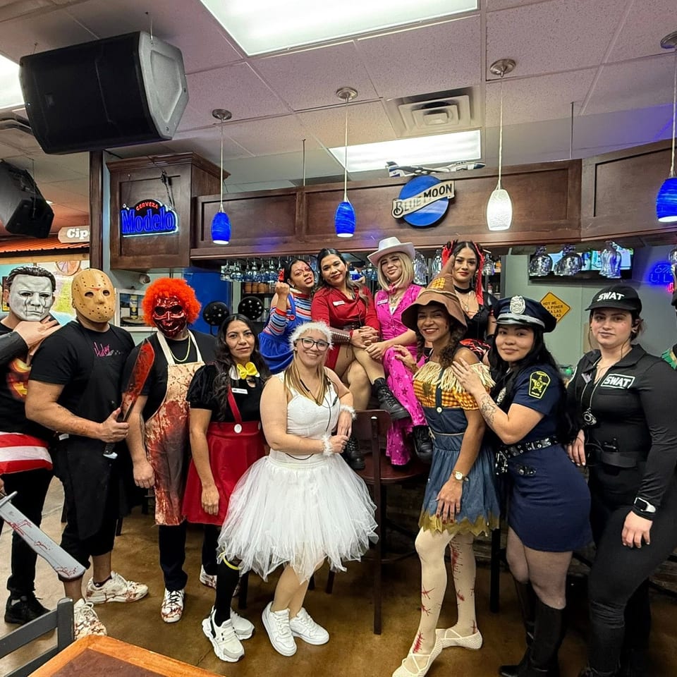 Group of people in colorful Halloween costumes posing together inside a lively bar beneath Modelo and Blue Moon signs.