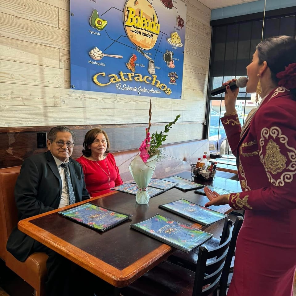 Restaurant guests listen to a singer in an ornate burgundy outfit beside a table with menus and flowers.