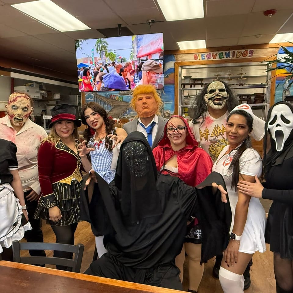 Restaurant staff and guests pose in colorful Halloween costumes beneath a “Bienvenidos” sign.