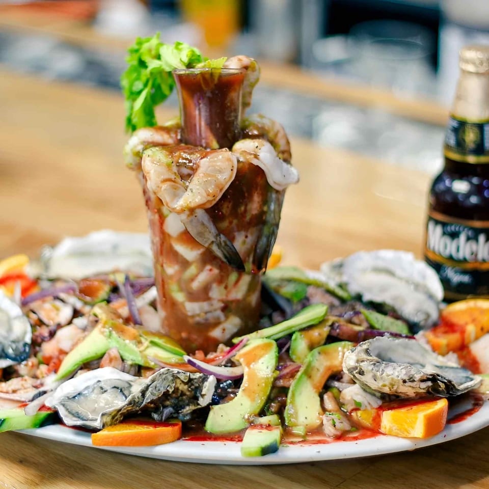 Seafood platter featuring shrimp cocktail, oysters, mussels, vegetables, citrus wedges, and a bottle of sauce.
