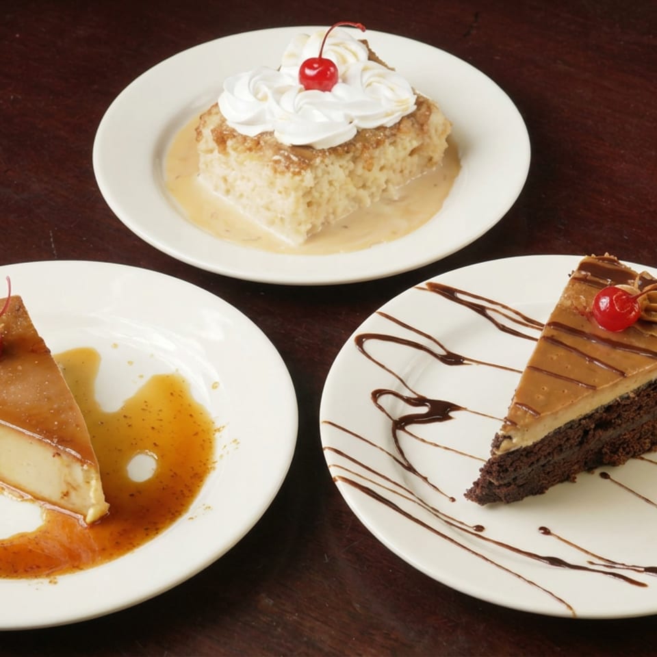 Three plated desserts—tres leches, flan, and chocolate cake—topped with whipped cream or cherries on a dark wooden table.